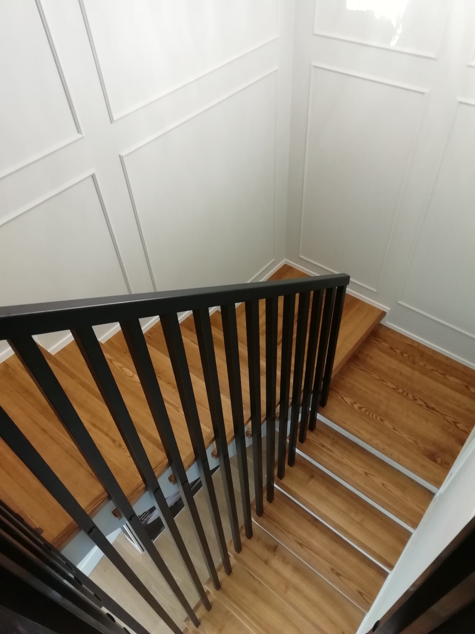 Kątowy widok z góry na drewniane schody z czarną, nowoczesną balustradą. Białe ściany z dekoracyjnymi listwami. Jasne, naturalne oświetlenie.