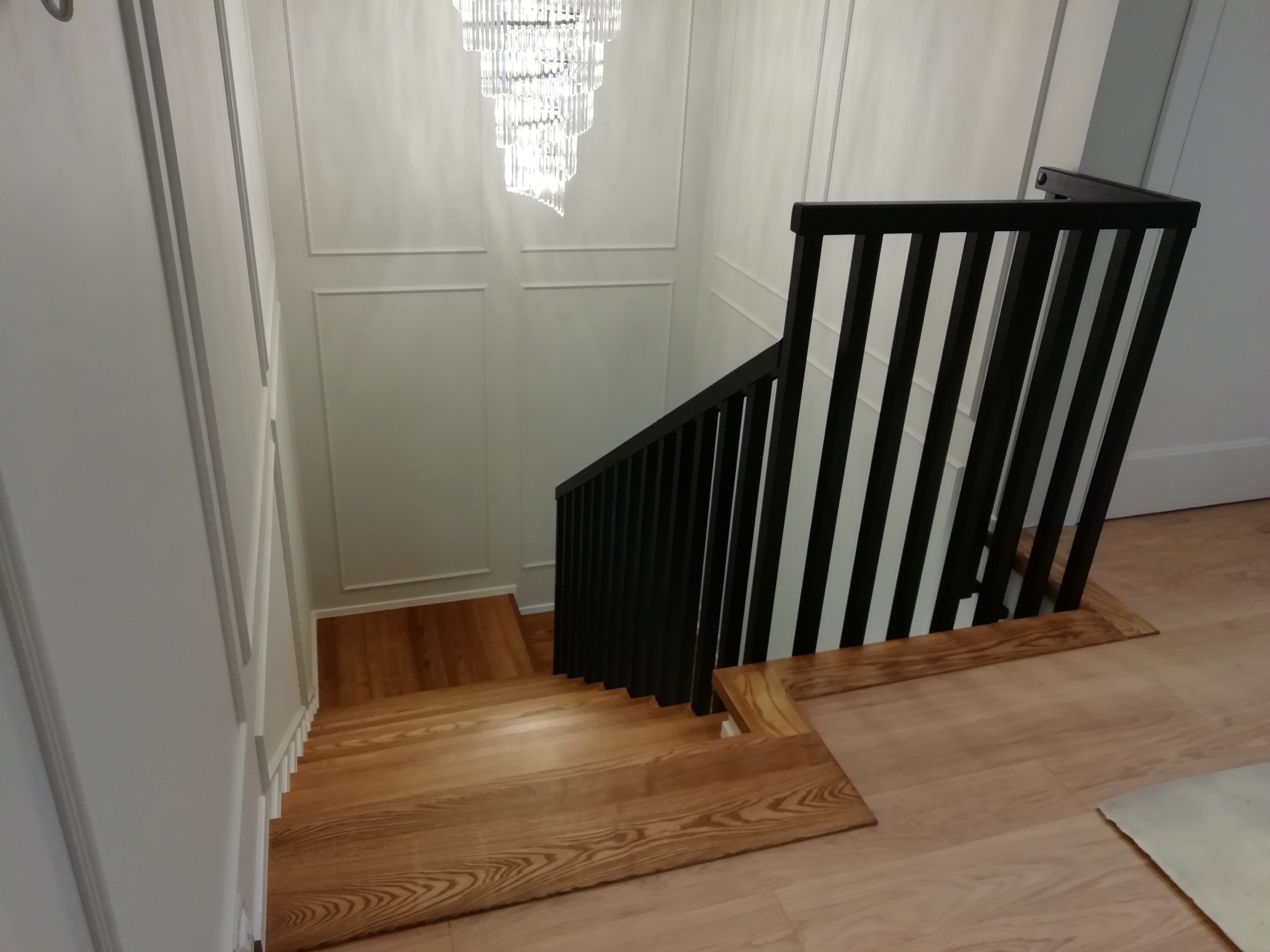 Drewniane schody z czarną, pionową balustradą w nowoczesnym wnętrzu z dekoracyjną ścianą i kryształowym żyrandolem, widok z góry.