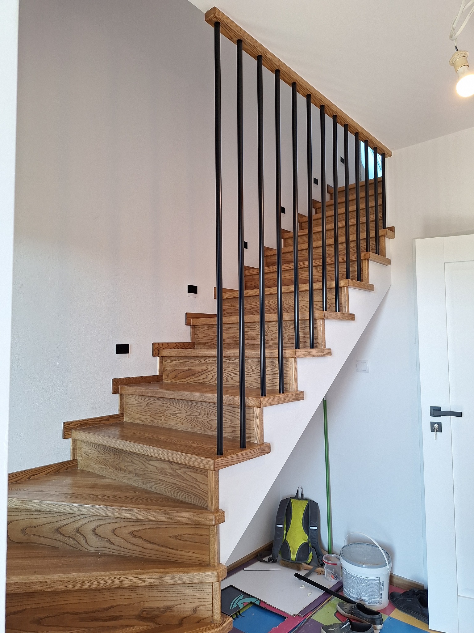 Jasne, drewniane schody z czarnymi, pionowymi balustradami, widziane z dołu. U dołu widoczne akcesoria malarskie i plecak, co sugeruje prace wykończeniowe.