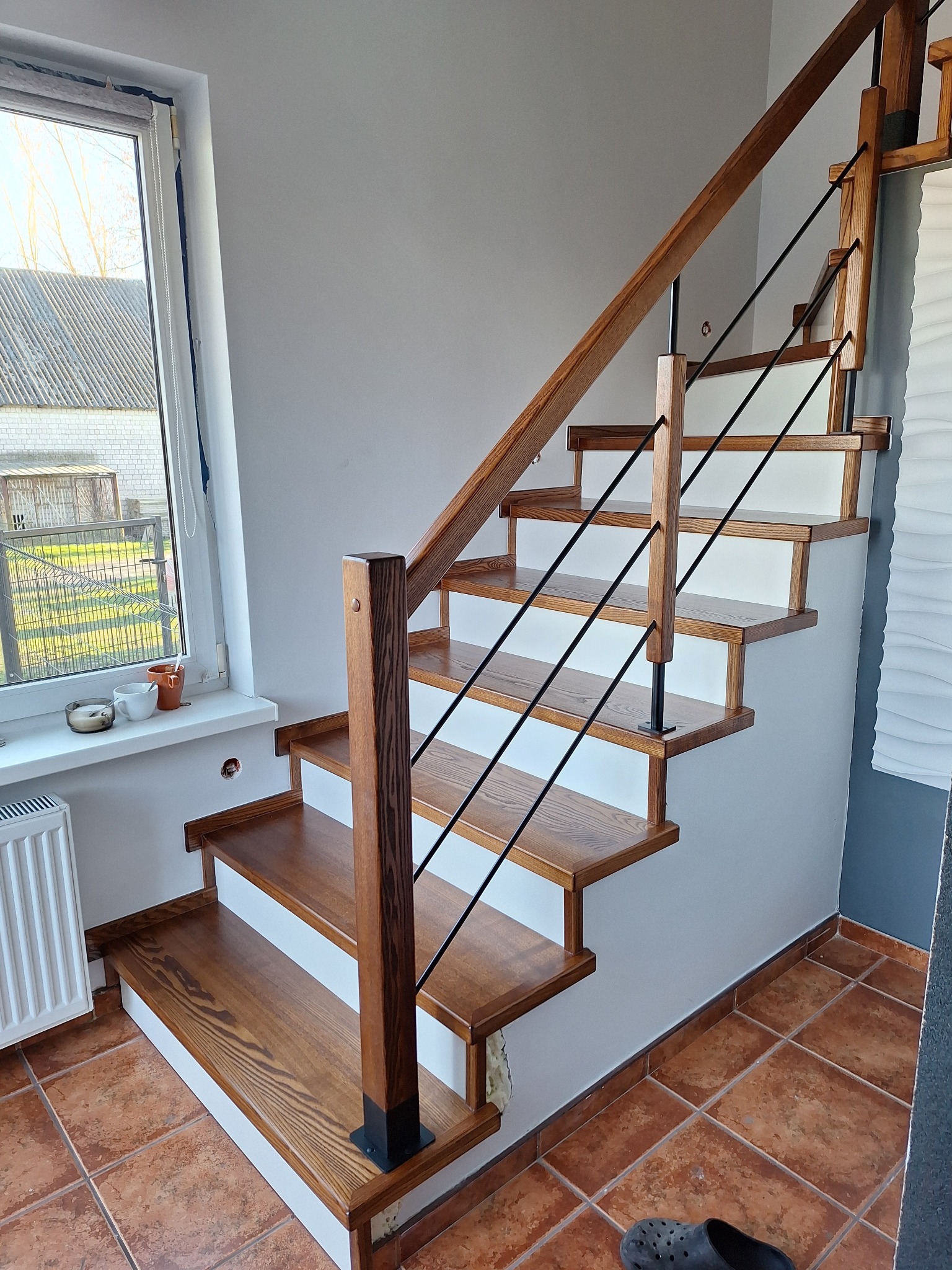 Nowoczesne schody drewniane z czarnymi, metalowymi balustradami i białymi podstopnicami, oświetlone naturalnym światłem z okna. Widok z dołu na konstrukcję.