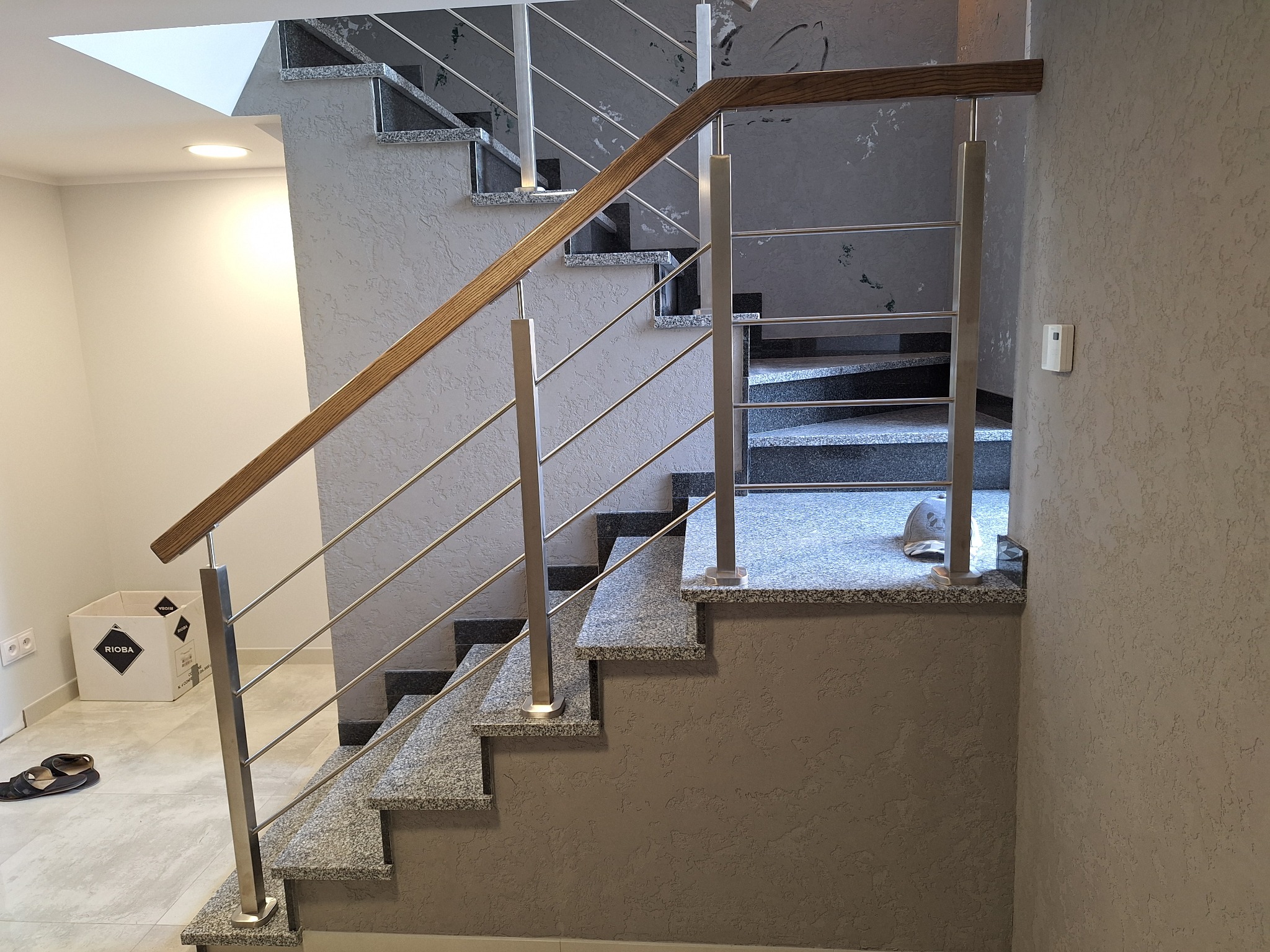 Granitowe schody z balustradą: drewniana poręcz i stalowe słupki. Widok z dołu, szare ściany z fakturą, pudełko i klapki w tle. Nowoczesny design.