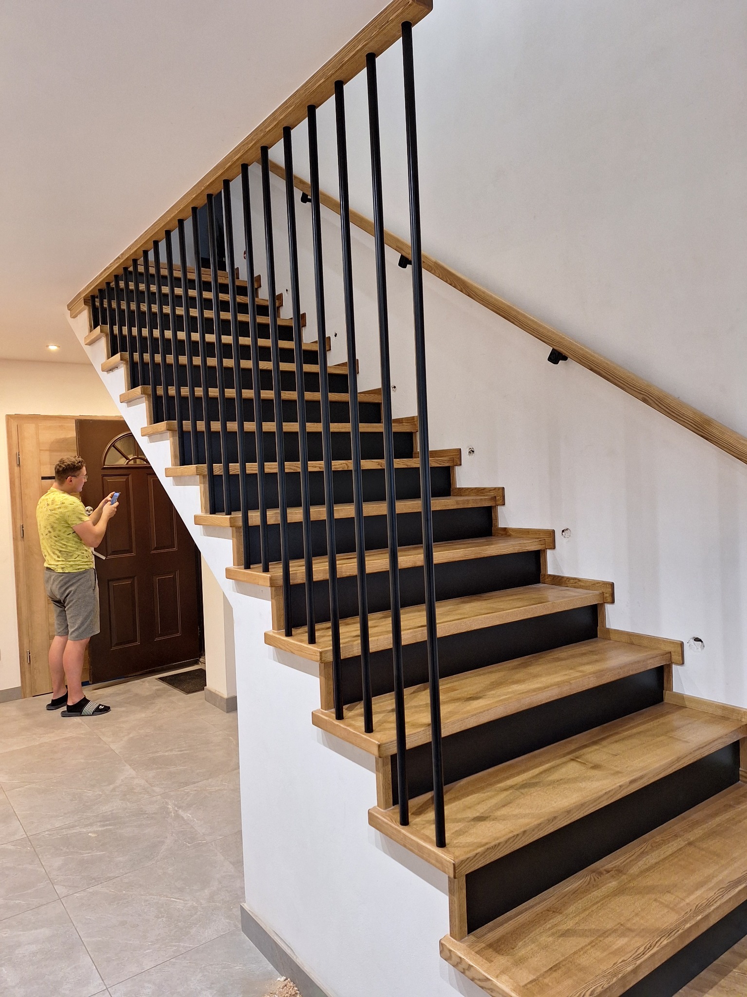 Nowoczesne schody drewniane z czarnymi, metalowymi balustradami i jasną poręczą. Mężczyzna robi zdjęcie schodów telefonem. Minimalistyczny design, jasne wnętrze.
