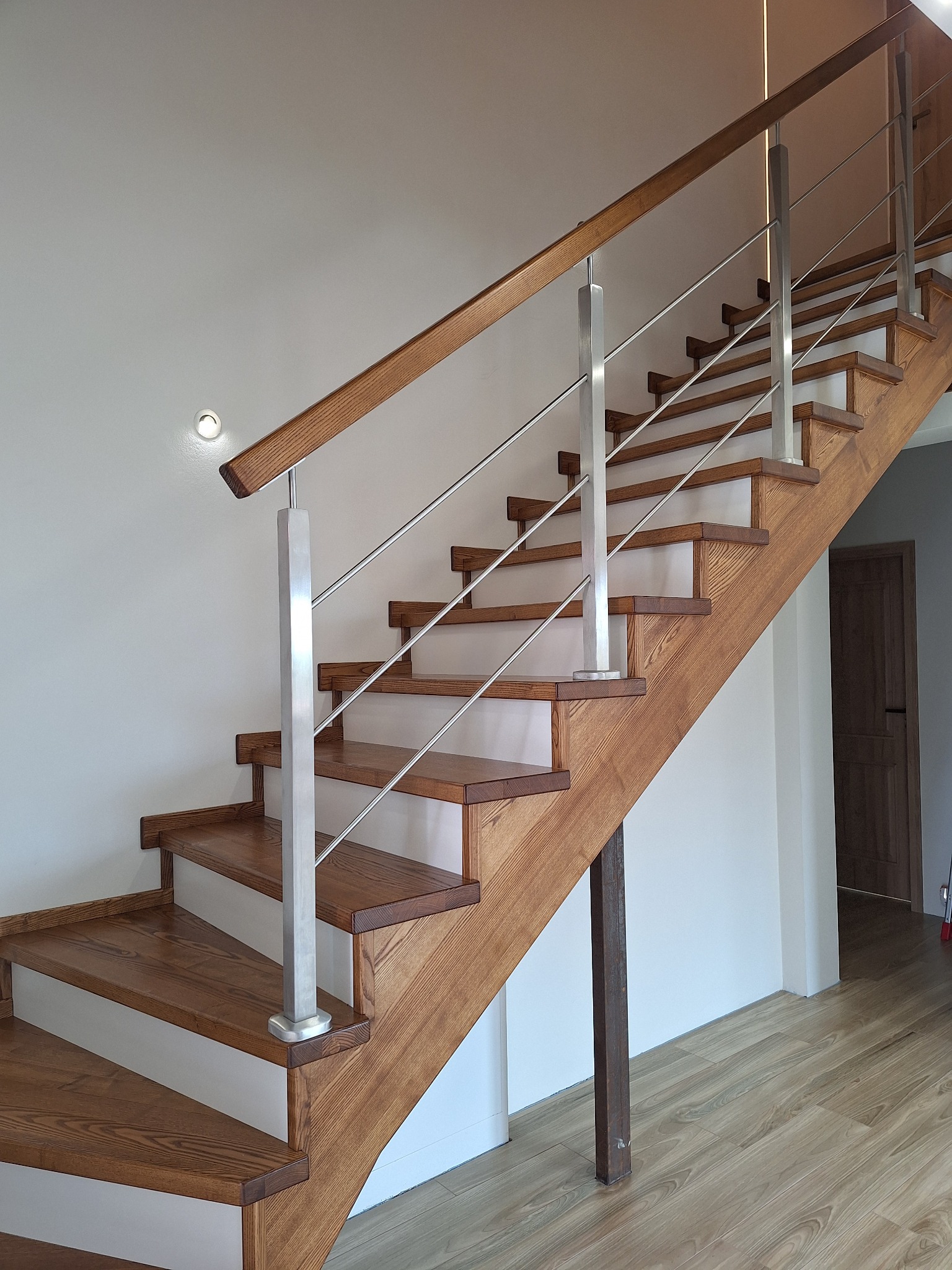 Drewniane schody z balustradą ze stali nierdzewnej, widok z dołu, oświetlenie punktowe, jasne ściany, drewniana podłoga, minimalistyczny design.