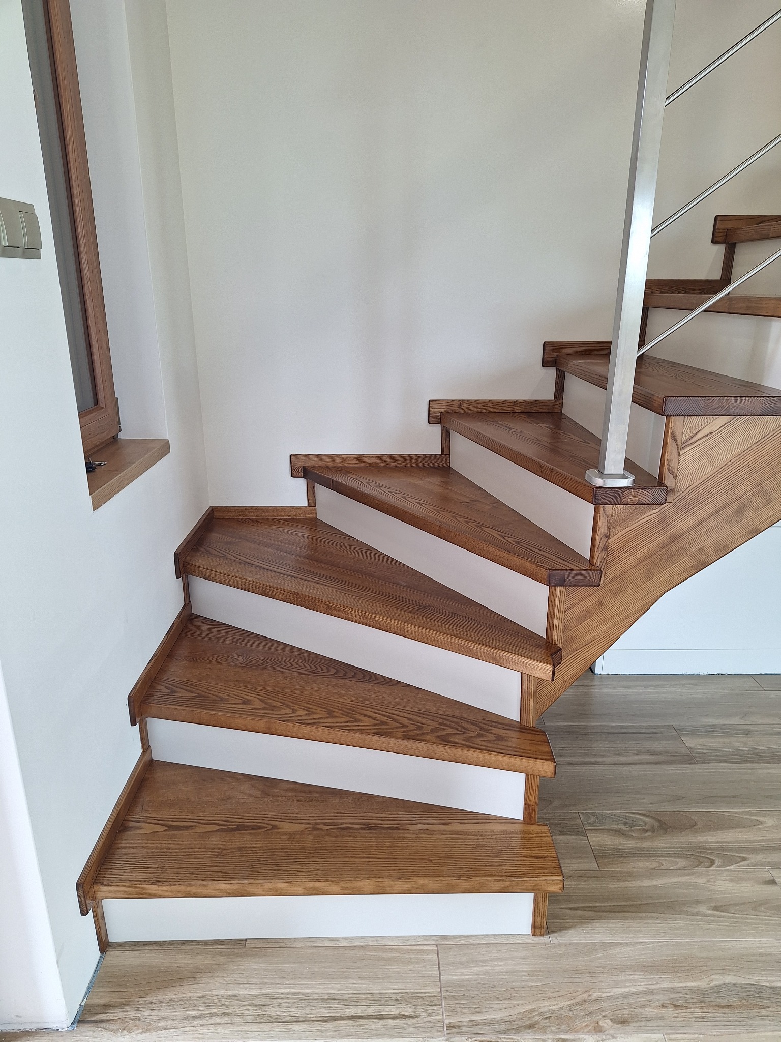 Drewniane schody z białymi podstopnicami i metalową balustradą, widoczne z dołu, z oknem w tle. Naturalne oświetlenie podkreśla strukturę drewna.