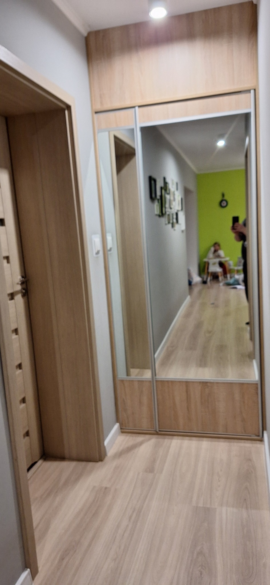Wnętrze przedpokoju z lustrzaną szafą przesuwną w odcieniach drewna, odbijającą dalszą część mieszkania z zieloną ścianą i osobą siedzącą przy stole.