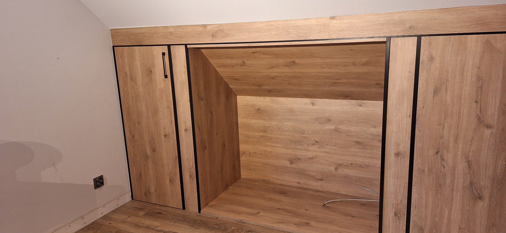 Zabudowa wnęki ze skosem z drewnopodobnej płyty w odcieniach brązu, z czarnymi obramowaniami, wykonana w miejscowości Wieniec. Drzwi przesuwne.