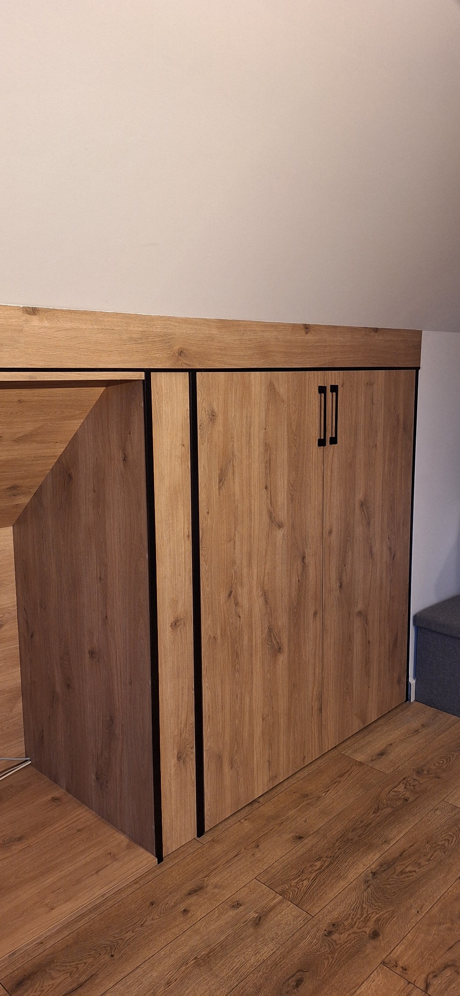 Drewniane schody z zabudową szafy pod skosem, wykończone w odcieniach dębu, z czarnymi uchwytami i obramowaniami, na tle podłogi z paneli.