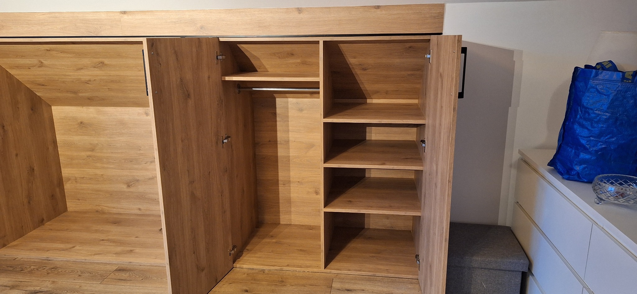 Otwarta, drewniana szafa z półkami i drążkiem na ubrania, widoczna faktura drewna, z szarą pufą i niebieską torbą w tle. Ujęcie z bliska, wnętrze szafy dobrze oświetlone.