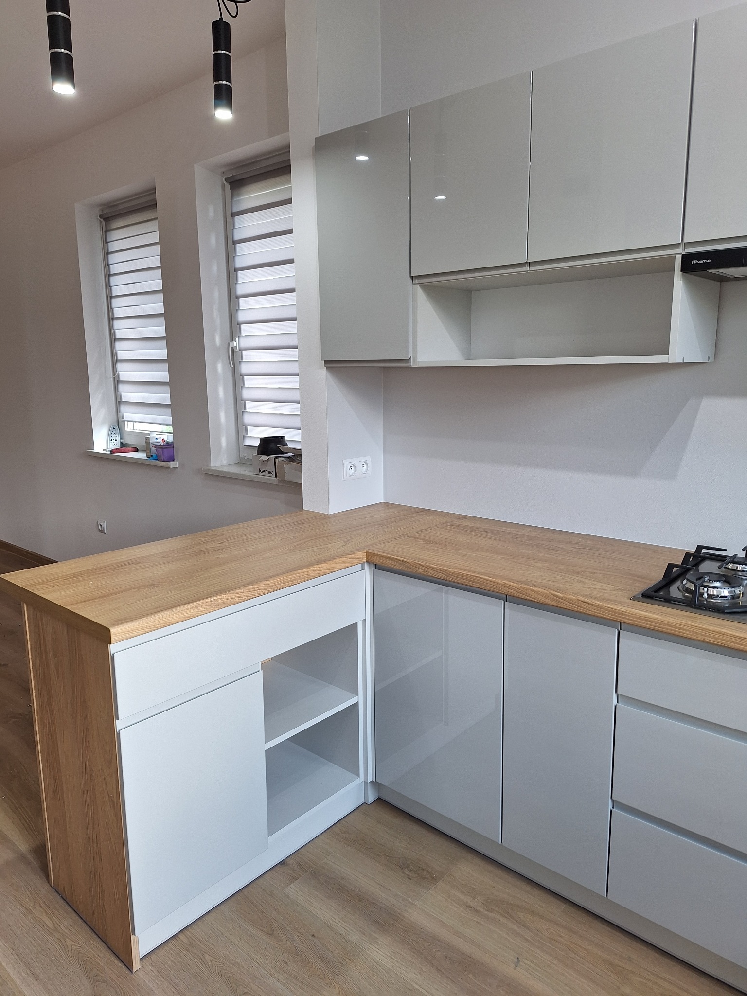 Nowoczesna kuchnia z szarymi, lakierowanymi frontami i drewnianym blatem. Widoczne okna z żaluzjami i minimalistyczne oświetlenie sufitowe.