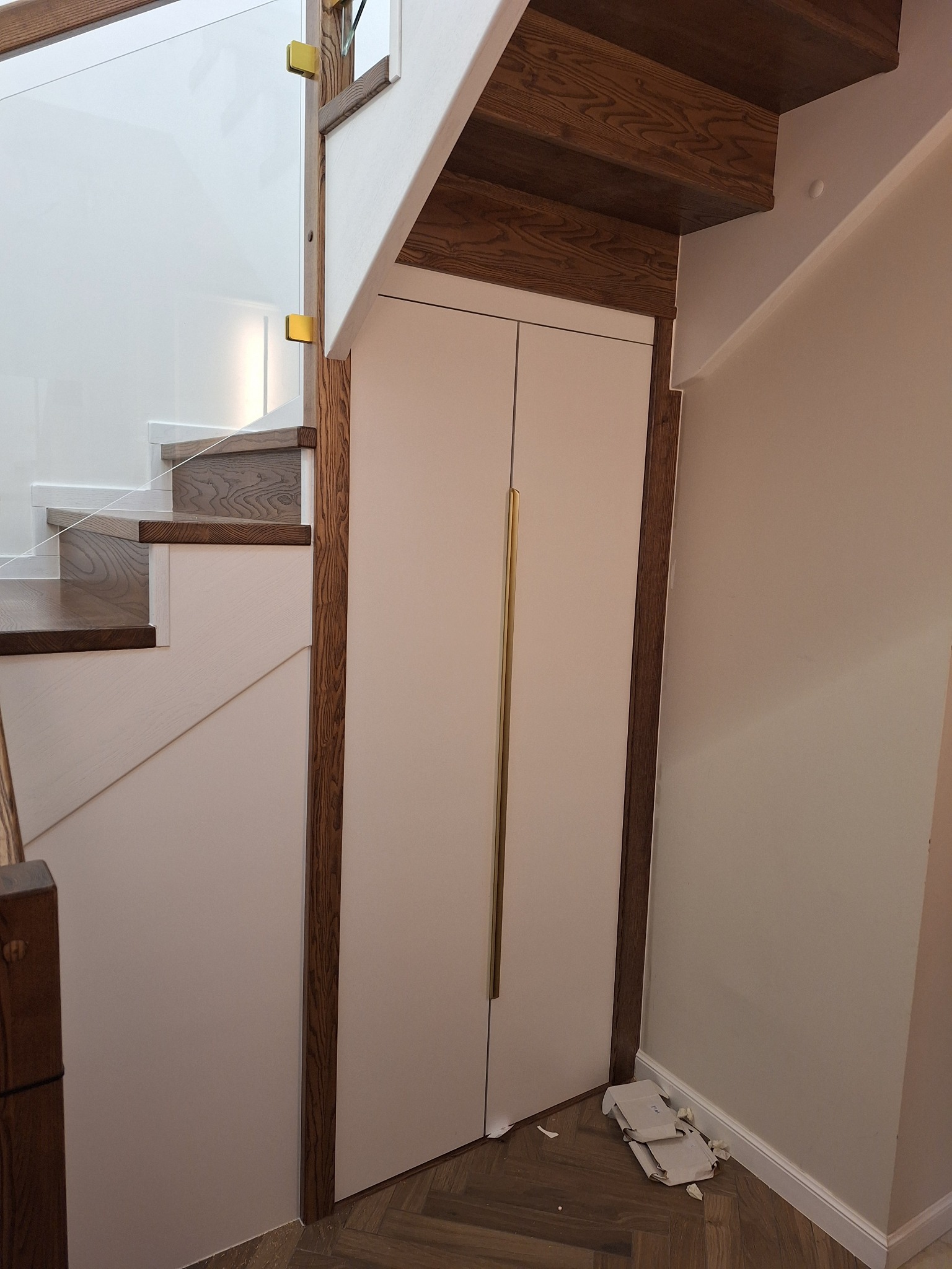 Nowoczesne schody z drewnianymi stopniami i szklaną balustradą, zintegrowane z szafą pod schodami, wykończone w jasnych kolorach i drewnianych akcentach.