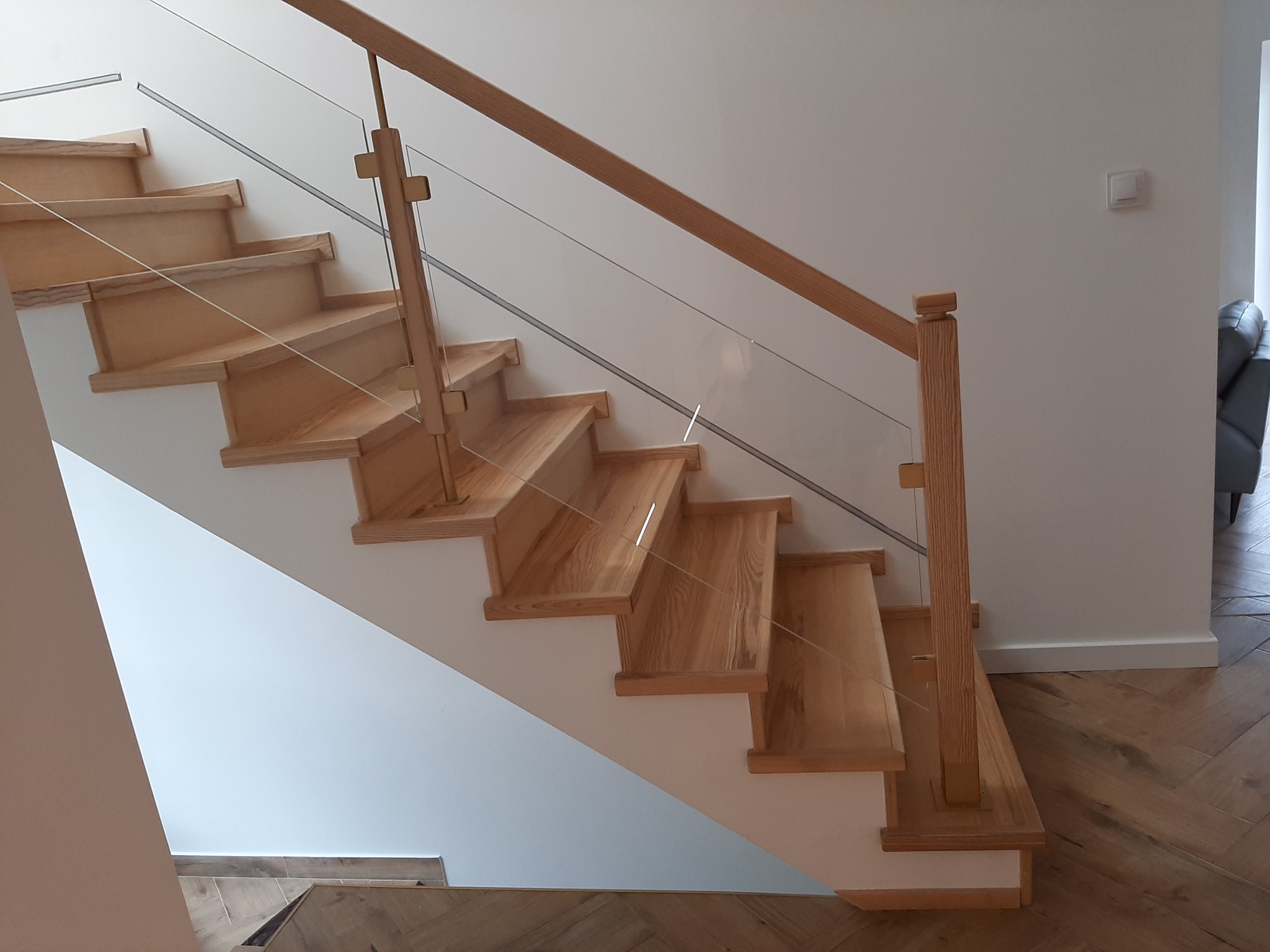 Jasne drewniane stopnie schodów z białymi podstopnicami, zabezpieczone szklaną balustradą z drewnianą poręczą, na podłodze parkiet ułożony w jodełkę.