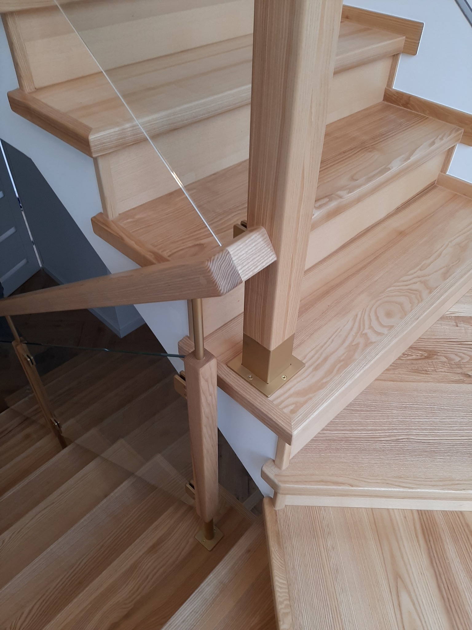 Jasne, drewniane stopnie z balustradą ze szkła i drewnianą poręczą, wspartą na złotych, metalowych elementach.