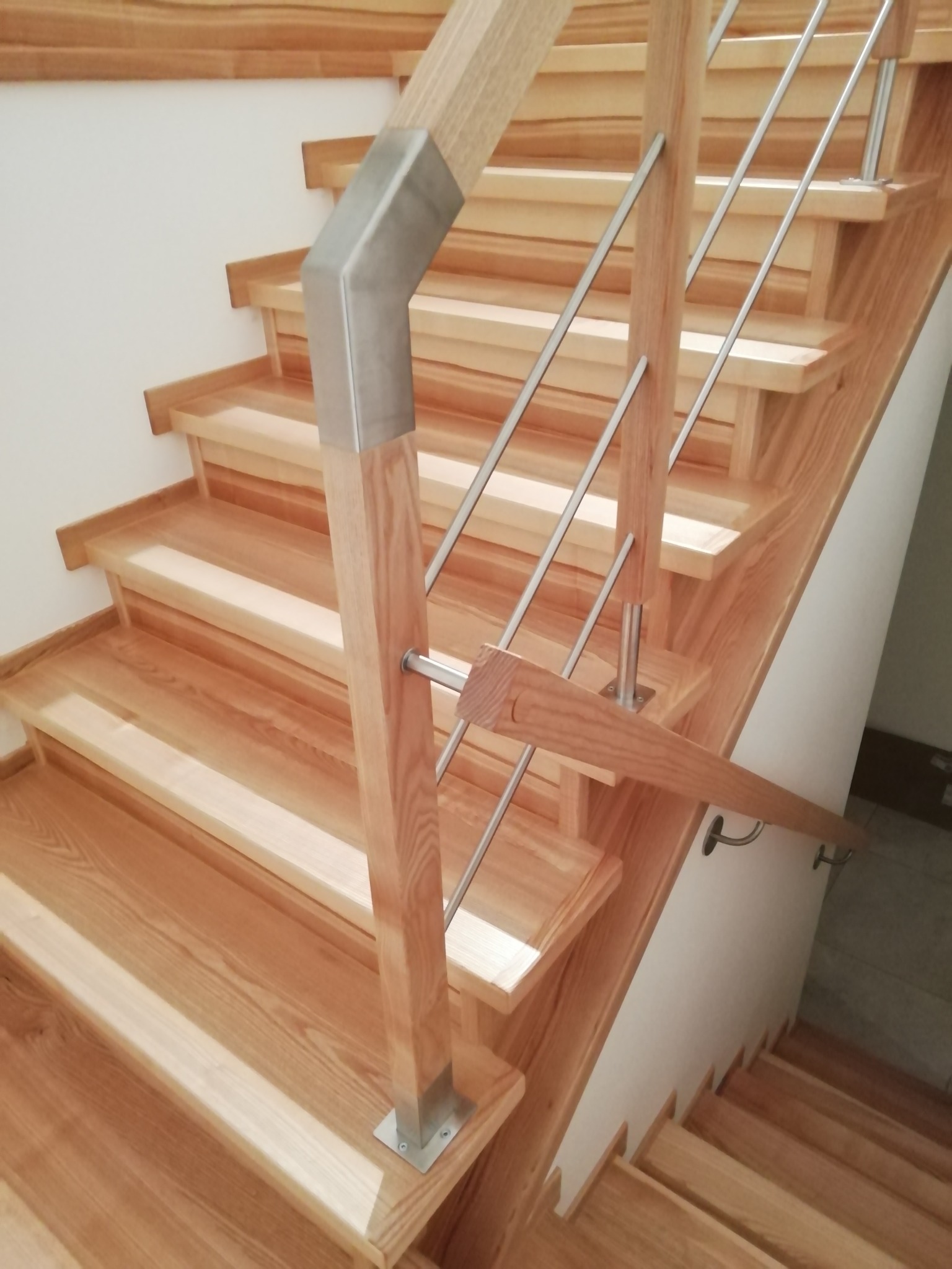 Jasne drewniane schody z balustradą z nierdzewnych prętów, zakończone metalowym łącznikiem, widok z dołu.