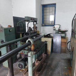 Zakład Ślusarski E. Bielik - Starodawna, zielona tokarka automatyczna z podajnikiem prętów, umieszczona w warsztacie z oknem w niebieskiej ramie i wagą szalkową w tle.