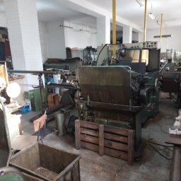 Zakład Ślusarski E. Bielik - Widok hali produkcyjnej z kilkoma maszynami do obróbki metalu, w tym tokarkami, widoczne pojemniki z wiórami, surowy beton na podłodze i białe ściany z cegły.