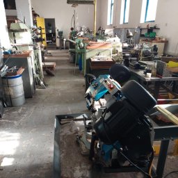 Zakład Ślusarski E. Bielik - Wnętrze warsztatu obróbki metali z widocznymi tokarkami i piłą taśmową do metalu na pierwszym planie, oświetlone naturalnym światłem wpadającym przez okna.