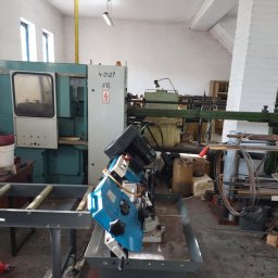 Zakład Ślusarski E. Bielik - Warsztatowa przestrzeń z widoczną piłą taśmową do metalu w kolorze niebieskim, ustawioną na metalowym stole, obok maszyny CNC i innych urządzeń, w tle białe ściany i sufit.