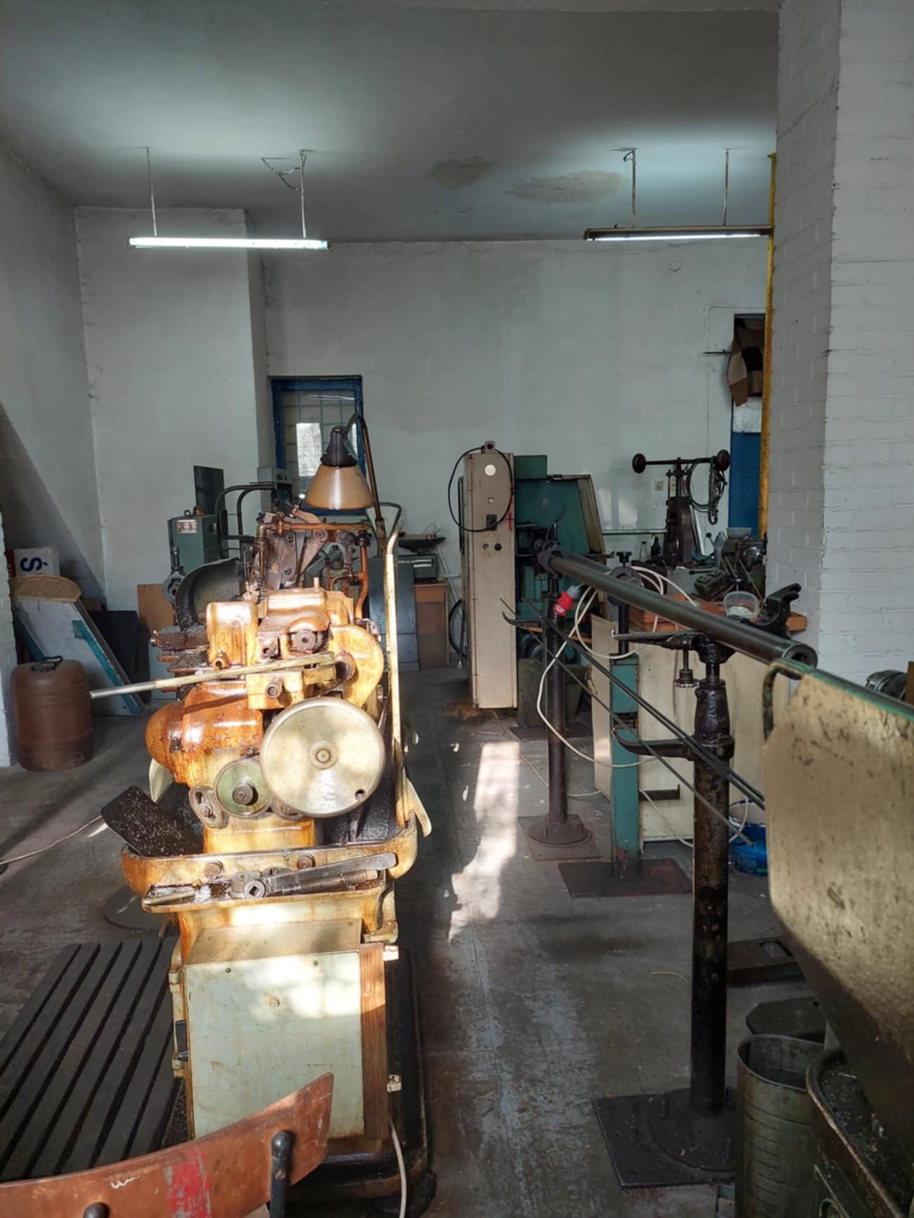 Stara, mocno zardzewiała maszyna do obróbki metalu ustawiona w warsztacie z widocznymi innymi urządzeniami i oświetleniem jarzeniowym.