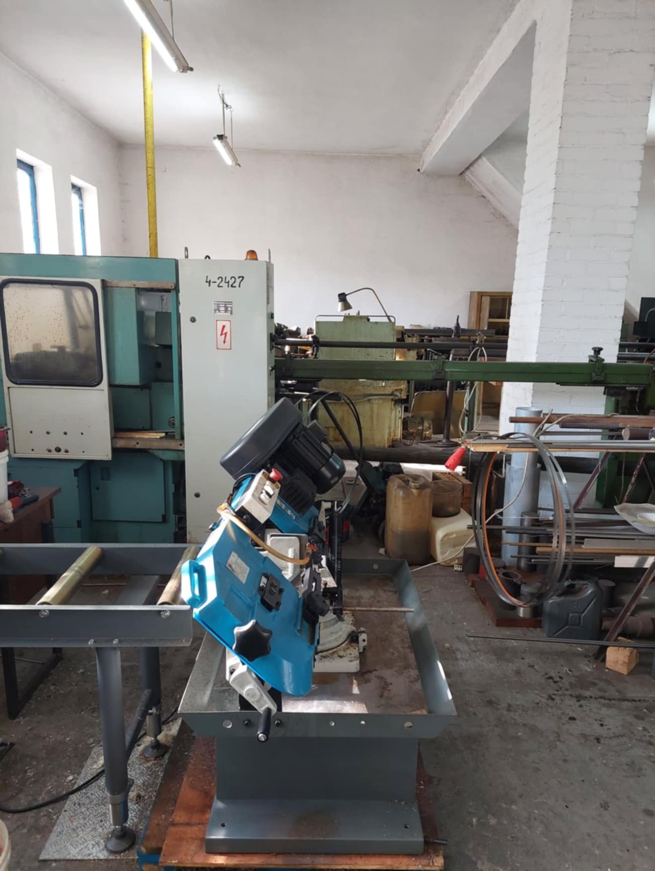 Warsztatowa piła taśmowa do metalu w hali produkcyjnej, obok starsza maszyna numeryczna z panelem sterowania, widoczne rury i materiały.