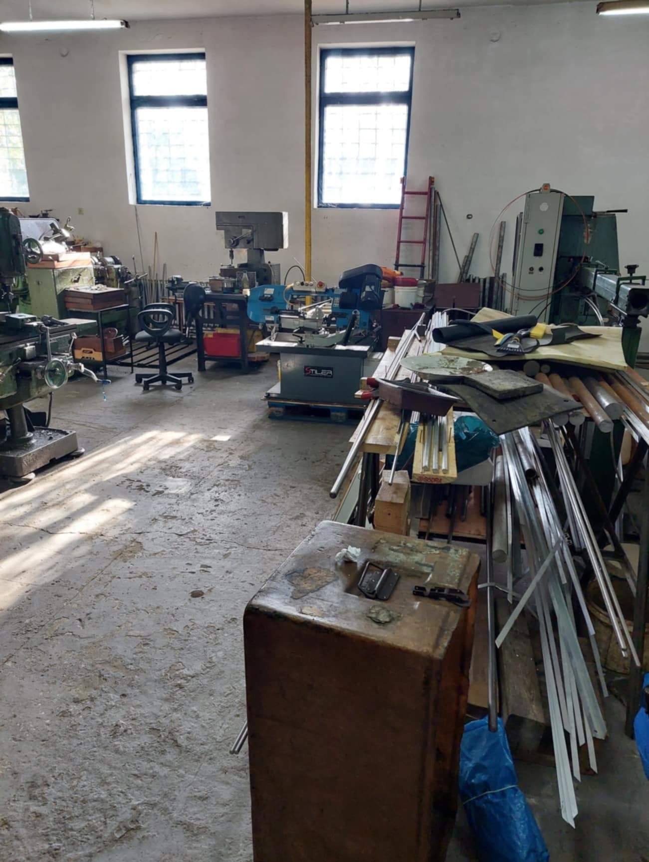 Wnętrze warsztatu z różnymi maszynami do obróbki metali, widoczne tokarki, frezarki i stół roboczy z materiałami. Na podłodze widoczne ślady użytkowania i rzucane cienie od światła wpadającego...