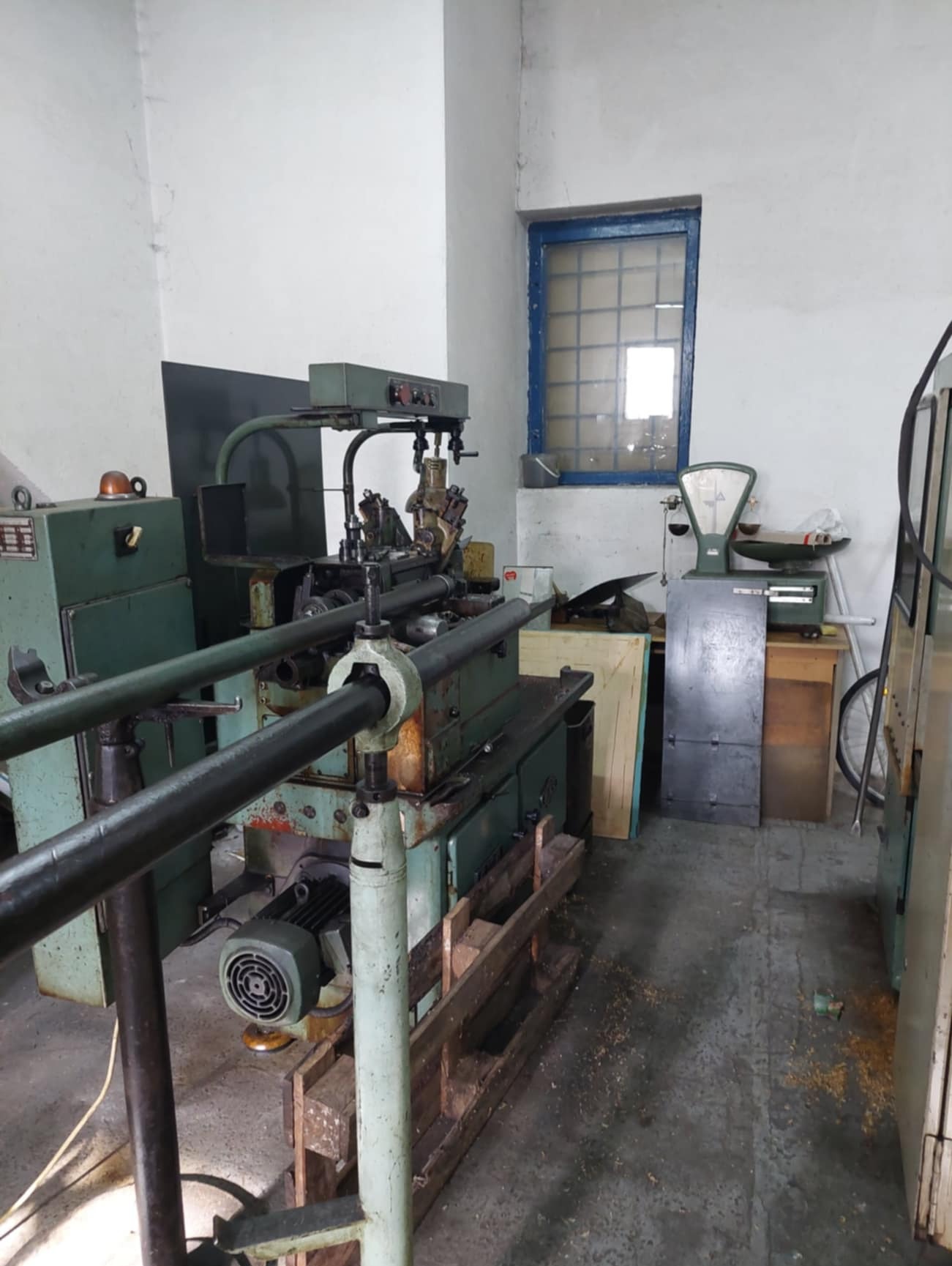 Starodawna, zielona tokarka automatyczna z podajnikiem prętów, umieszczona w warsztacie z oknem w niebieskiej ramie i wagą szalkową w tle.