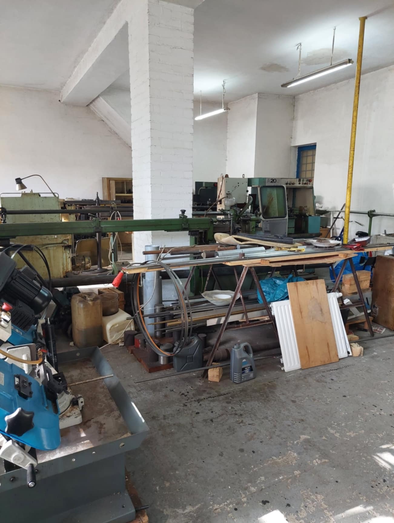 Warsztat obróbki metali: widok ogólny z widocznymi tokarkami, frezarkami i materiałami składowanymi na stołach roboczych, w tle kolumna pomalowana na biało.