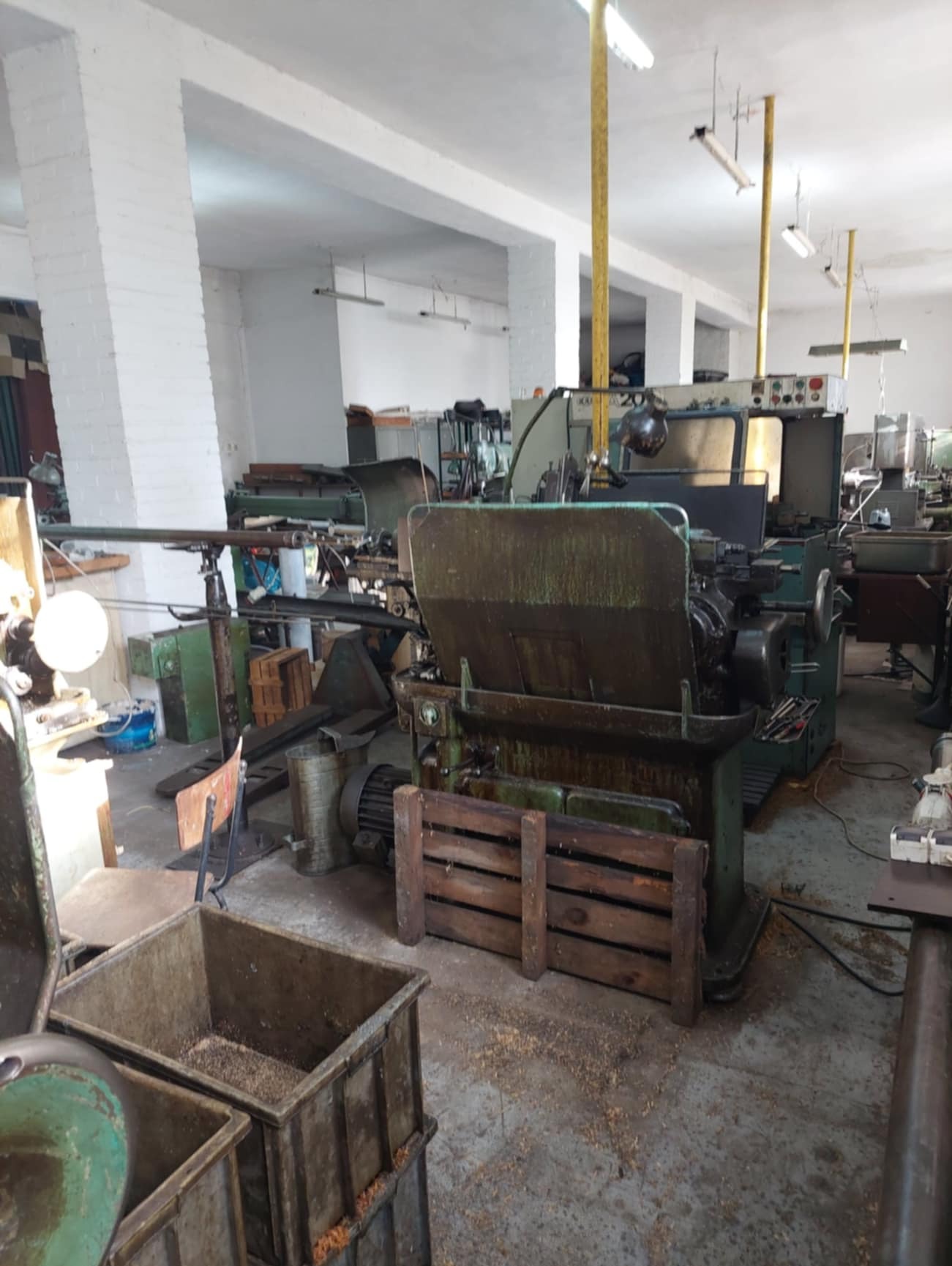 Widok hali produkcyjnej z kilkoma maszynami do obróbki metalu, w tym tokarkami, widoczne pojemniki z wiórami, surowy beton na podłodze i białe ściany z cegły.