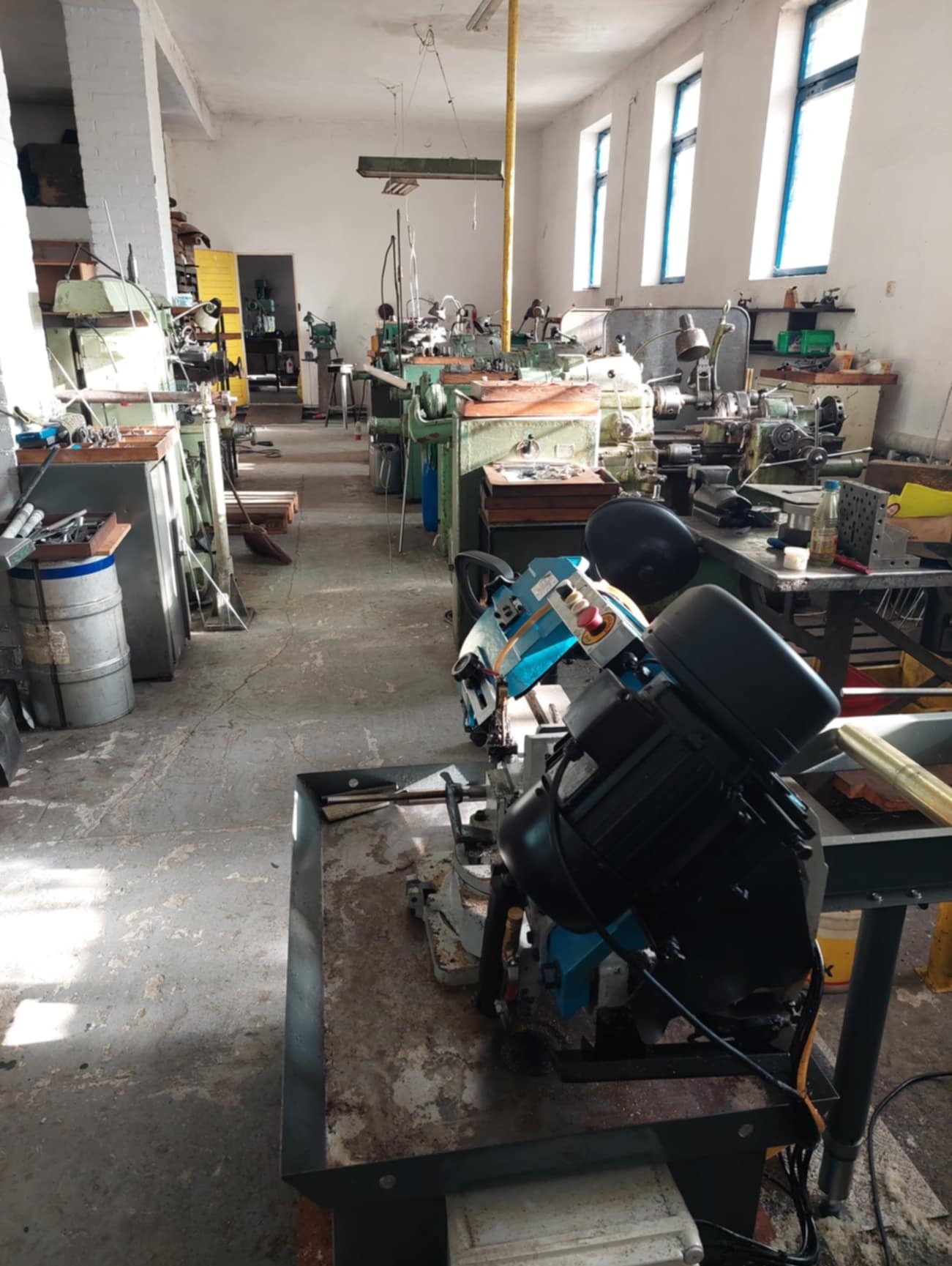 Wnętrze warsztatu obróbki metali z widocznymi tokarkami i piłą taśmową do metalu na pierwszym planie, oświetlone naturalnym światłem wpadającym przez okna.