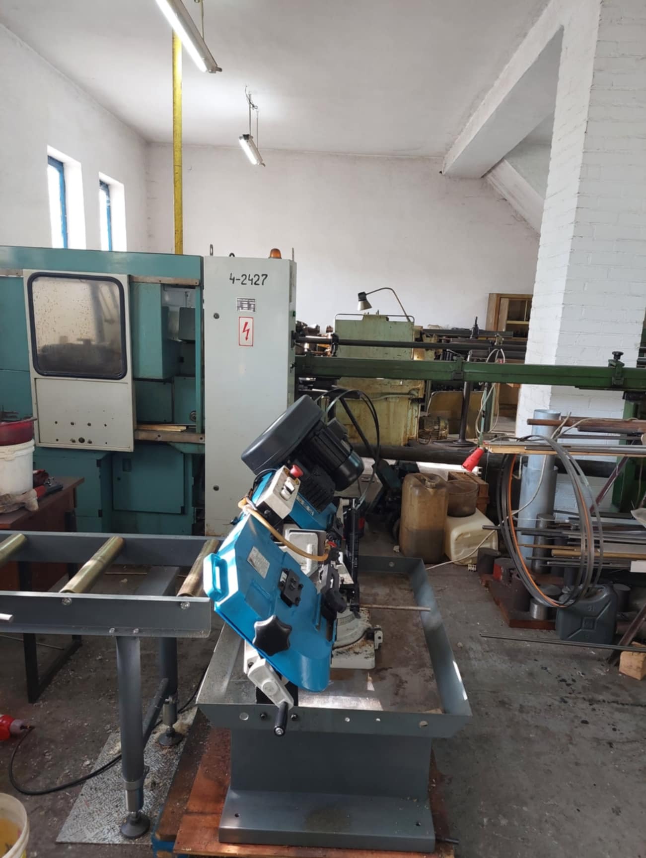 Warsztatowa przestrzeń z widoczną piłą taśmową do metalu w kolorze niebieskim, ustawioną na metalowym stole, obok maszyny CNC i innych urządzeń, w tle białe ściany i sufit.