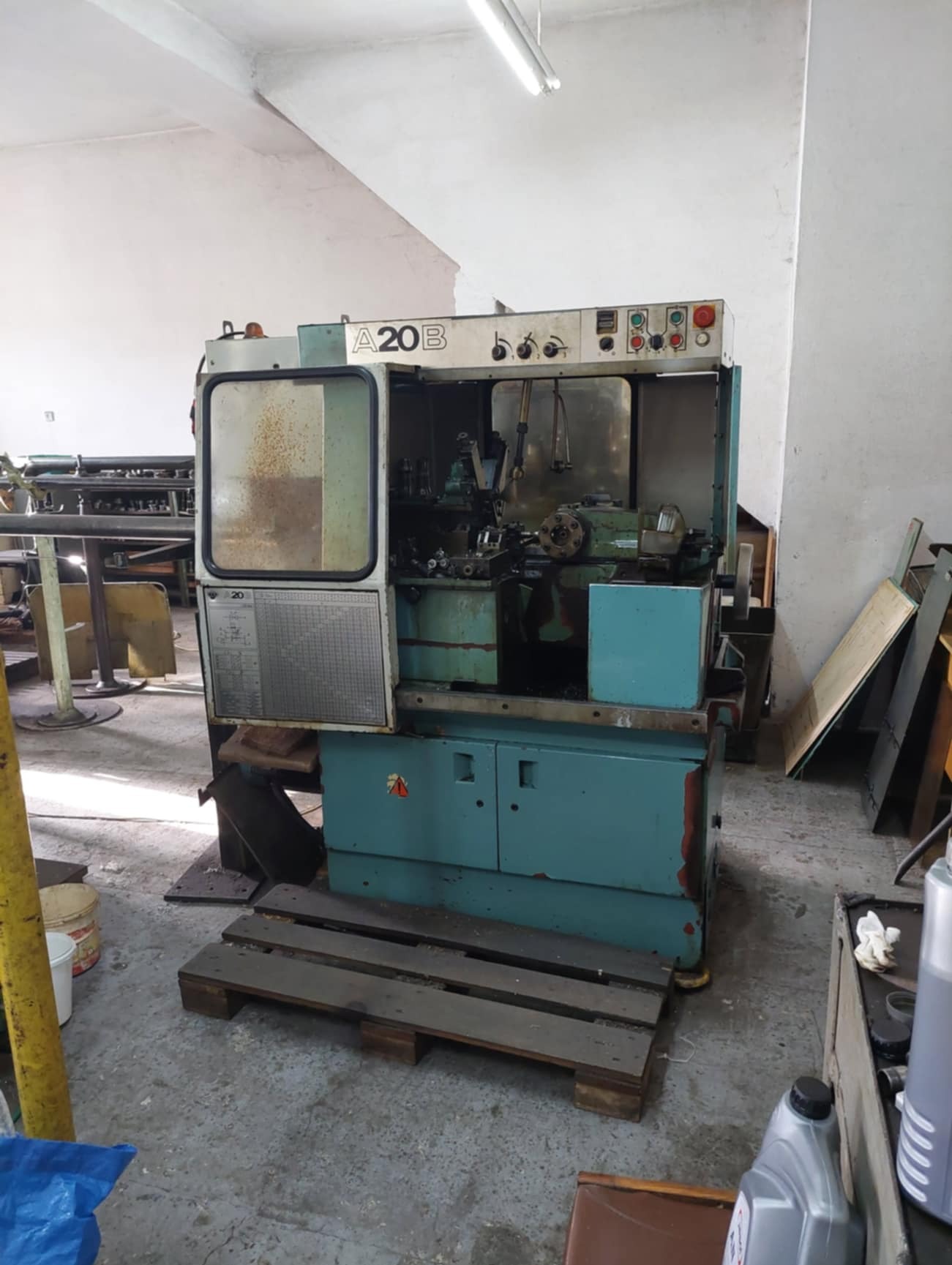 Używana, niebieska tokarka automatyczna A20B z otwartą osłoną, stojąca na drewnianej palecie w warsztacie.