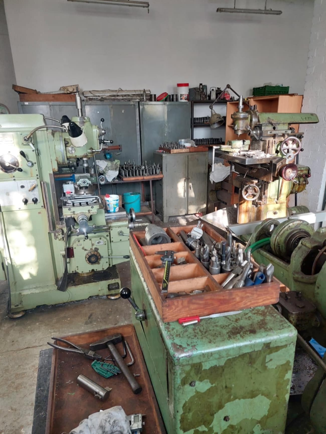 Wnętrze warsztatu z dwoma maszynami do obróbki metali w odcieniach zieleni, skrzynią z narzędziami, widoczne wiertła, suwmiarka, młotek i inne narzędzia na stole warsztatowym, w tle regały i szafki...
