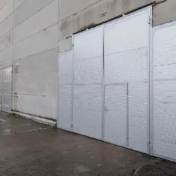 Duża, segmentowa, metalowa brama przemysłowa w kolorze srebrnym, z małym wejściem po prawej stronie, zamontowana w szarym, betonowym budynku przemysłowym. Widoczna mokra nawierzchnia przed budynkiem.