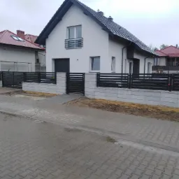 Nowoczesne, poziome, czarne panele ogrodzeniowe zamontowane na niskim murku z betonowych bloczków w kolorze szarym, na tle nowego domu z białą elewacją i szarym dachem, widok z poziomu ulicy.