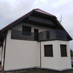 AMB-MET - Dwupiętrowy dom z beżową elewacją, ciemnobrązową stolarką okienną i poziomą balustradą balkonową w tym samym kolorze, widoczny z zewnątrz w pochmurny dzień.