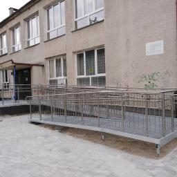 AMB-MET - Zewnętrzna rampa dla niepełnosprawnych z metalową konstrukcją i balustradami, prowadząca do wejścia budynku o szarej elewacji z widocznymi śladami użytkowania.