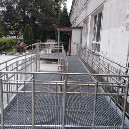 AMB-MET - Zewnętrzna rampa dla niepełnosprawnych z metalową kratownicą i balustradami ze stali nierdzewnej, prowadząca do budynku z widocznymi śladami użytkowania.
