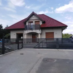 Nowoczesna posesja w Wołowie z grafitową, poziomą bramą wjazdową i ogrodzeniem panelowym, dopasowanymi do balustrady balkonowej w domu z czerwoną dachówką.