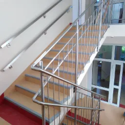 Wewnętrzne schody z metalową balustradą nierdzewną, widok z góry, jasne wnętrze budynku z czerwonymi akcentami na podłodze i szarymi krawędziami stopni.