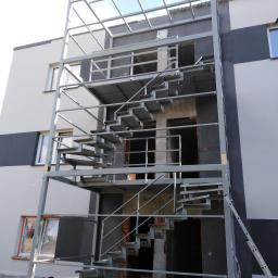 AMB-MET - Zewnętrzne, metalowe schody ewakuacyjne w trakcie montażu na ścianie budynku, widoczne z dołu pod kątem, z pracownikiem w odblaskowej kamizelce w tle.