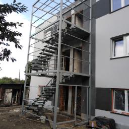 AMB-MET - Zewnętrzne schody metalowe z ramą ochronną, przymocowane do boku budynku w trakcie budowy, widoczne materiały budowlane i bałagan na ziemi.
