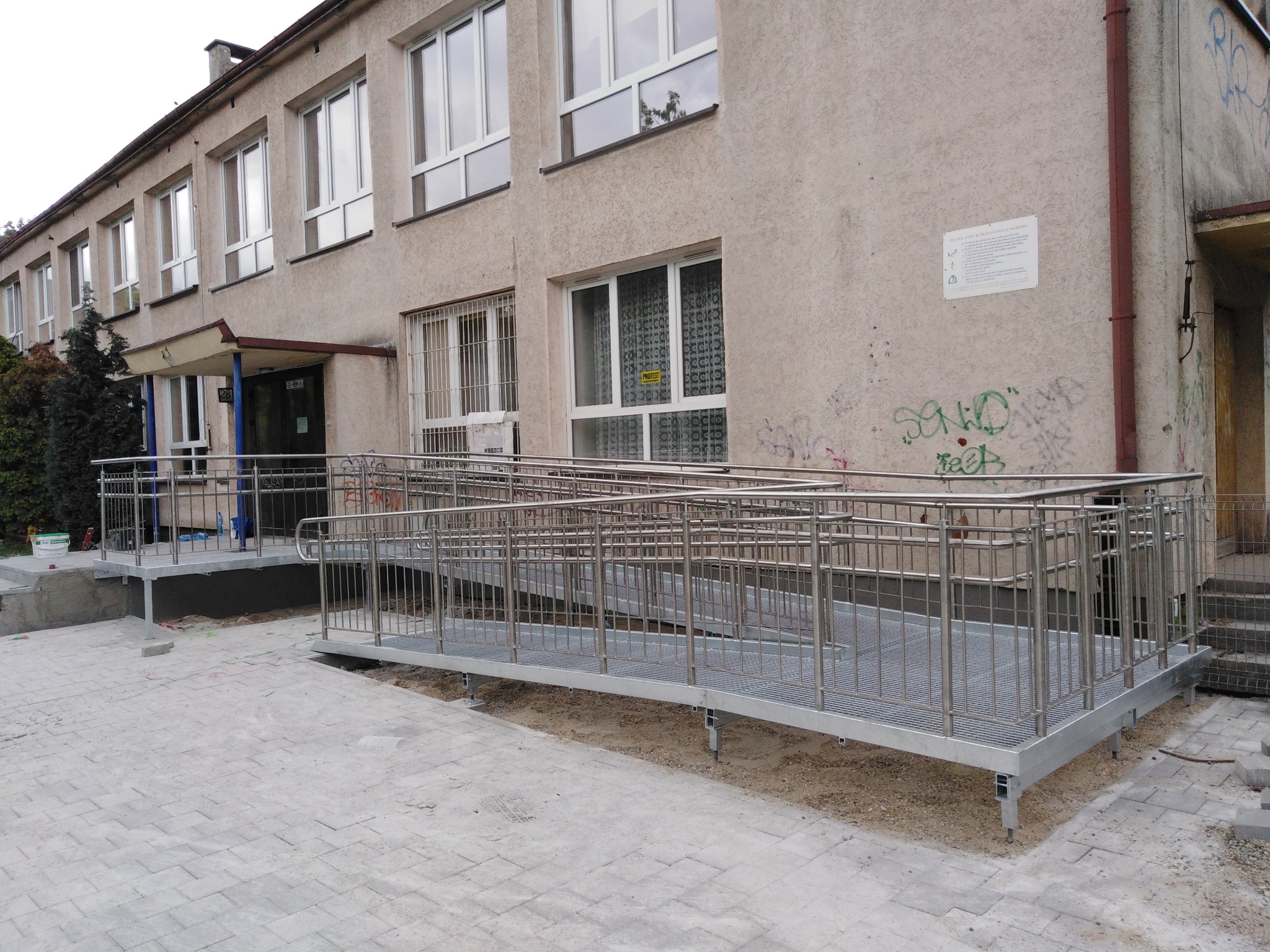 Zewnętrzna rampa dla niepełnosprawnych z metalową konstrukcją i balustradami, prowadząca do wejścia budynku o szarej elewacji z widocznymi śladami użytkowania.