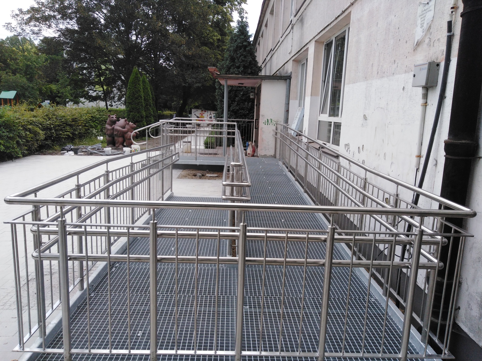 Zewnętrzna rampa dla niepełnosprawnych z metalową kratownicą i balustradami ze stali nierdzewnej, prowadząca do budynku z widocznymi śladami użytkowania.