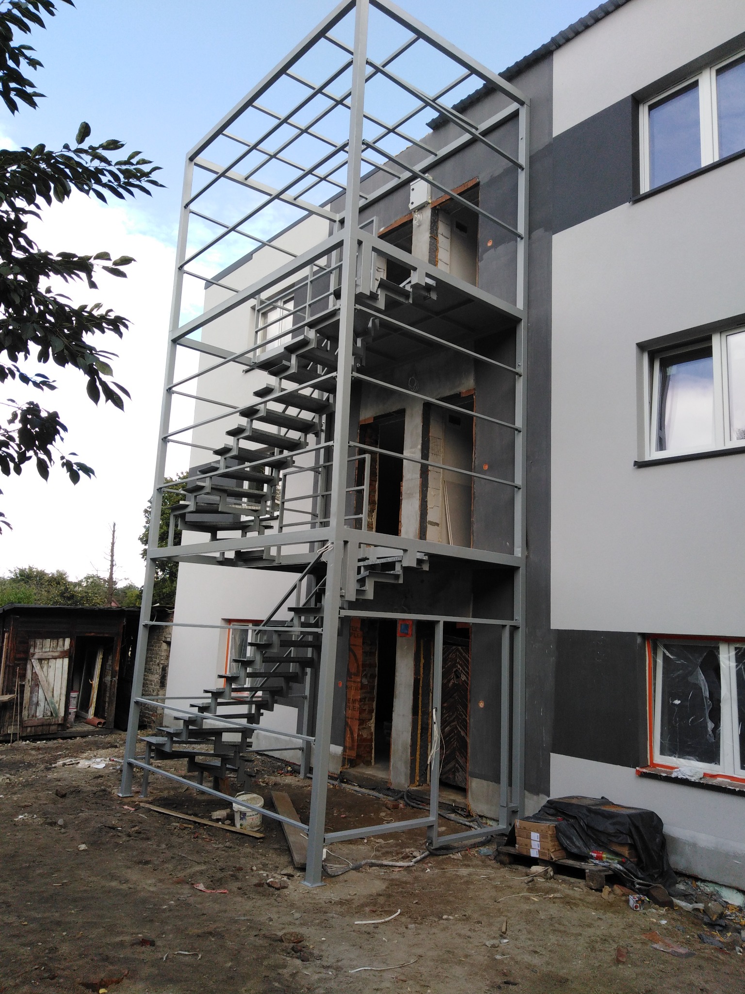Zewnętrzne schody metalowe z ramą ochronną, przymocowane do boku budynku w trakcie budowy, widoczne materiały budowlane i bałagan na ziemi.