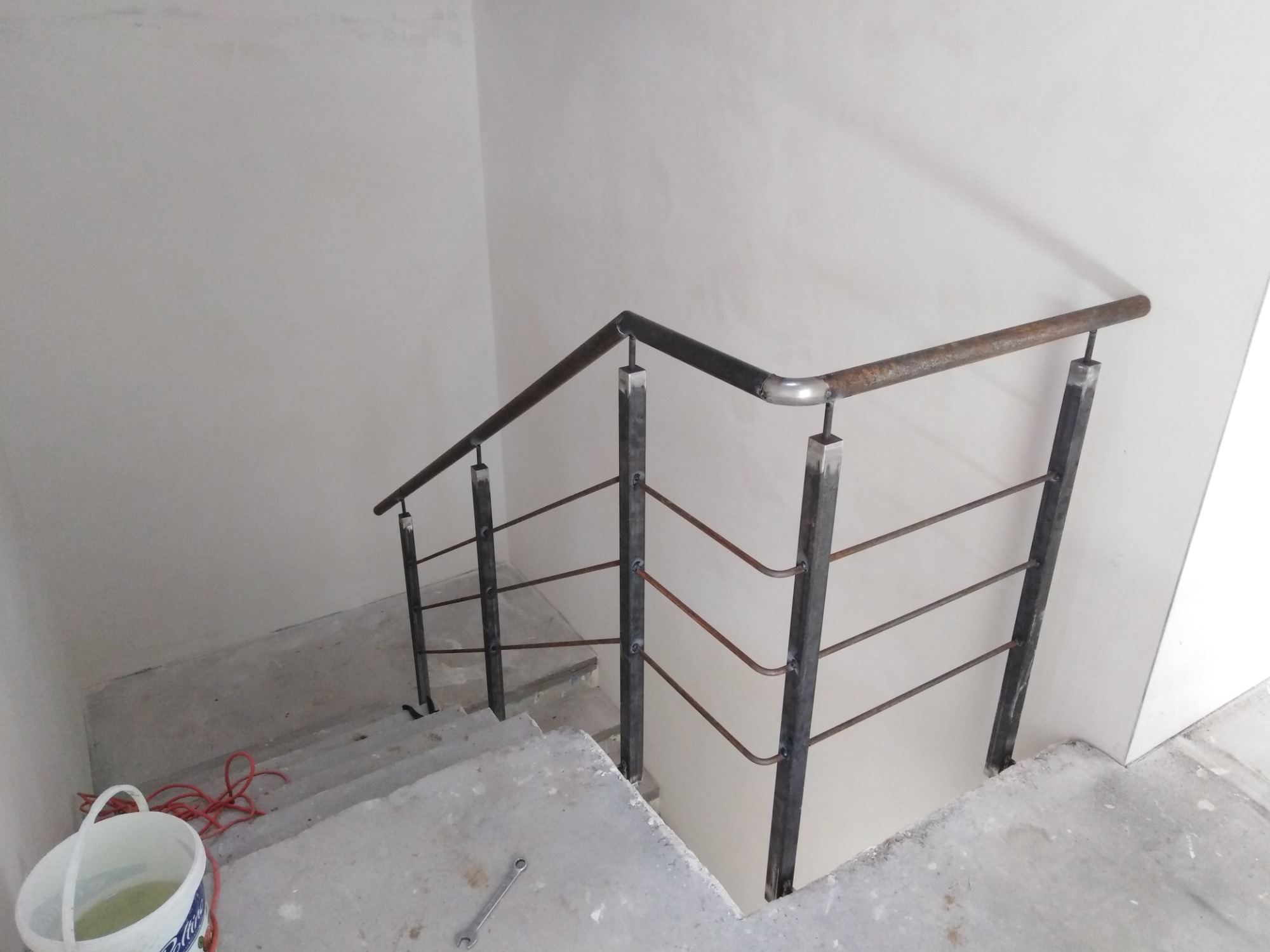 Surowe, metalowe balustrady schodowe w trakcie montażu, widoczne ślady spawania, betonowe schody w stanie surowym, wiadro z wodą i narzędzie na podłodze.