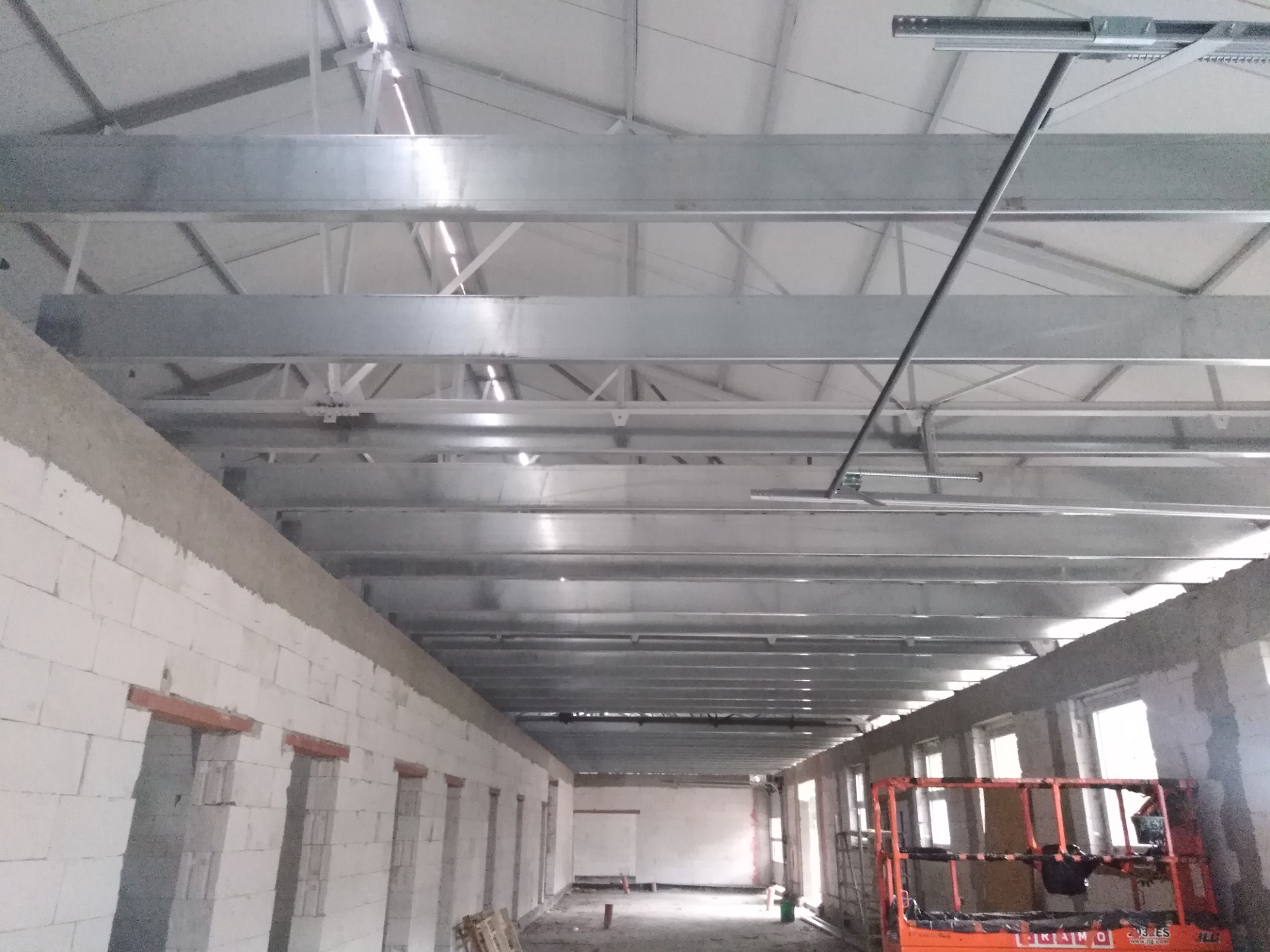 Wnętrze hali produkcyjnej w trakcie budowy, widoczne surowe ściany z betonu komórkowego, stalowa konstrukcja dachu i podnośnik koszowy.