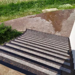 Zewnętrzne schody z brązowo-szarego kamienia, prowadzące na taras z wzorem, widok z góry, w tle trawa i drzewa.