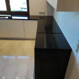 Czarny, błyszczący blat kuchenny z wbudowaną płytą grzewczą, widoczny pod kątem, z białymi szafkami w tle i oknem z widokiem na zewnątrz.