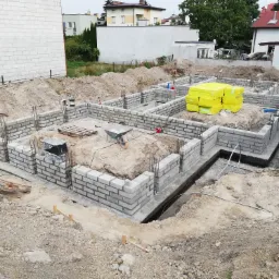 Wykopane i częściowo wylane fundamenty pod budynek mieszkalny, widoczne bloczki betonowe, wystające pręty zbrojeniowe, żółte paczki styropianu oraz taczka z piaskiem.