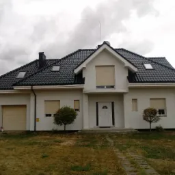 Nowoczesny dom jednorodzinny z czarną dachówką, białymi ścianami i zamkniętymi roletami w oknach, widziany z perspektywy trawnika w pochmurny dzień.