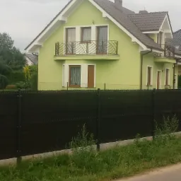 Jasnozielony dom jednorodzinny z brązowym dachem i balkonami z ozdobnymi balustradami, widoczny zza czarnego ogrodzenia panelowego.